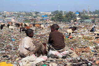 An photograph of two boys sitting on a dumpsite with grazing cows and the city in the background.