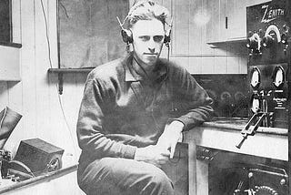 A picture from what appears to be the early 20th century of a man sitting at a desk with headphones on listening to a ham radio.