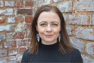 A woman with brown hair, wearing a black turtleneck, against a background of bricks.