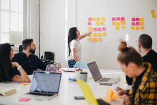 Group of professionals sitting around a conference table watching a long haired woman move stickies around a whiteboard.