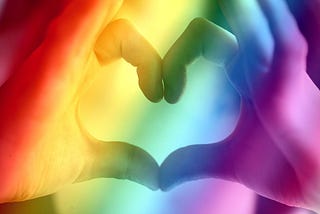 A person’s hands making the shape of a heart. The photo has a rainbow filter.