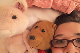 A selfie of Lyndsey laying on a bed that has pink sheets. She’s with a brown bear and a pink pig that blends into the sheets.