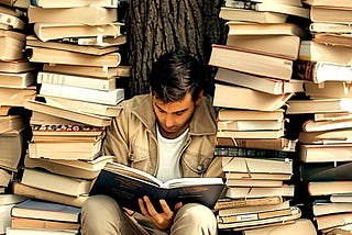 A writer is sitting against a tree looking at a book. He is surrounded by stacks of books. Image produced using Midjourney