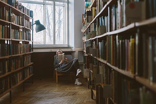 A lady reading on a chair in between two large book shelves