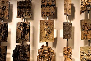 This image displays a collection of Benin bronzes. These intricate metal plaques depict various figures in traditional attire, often engaged in ceremonial or ritualistic activities. The bronzes showcase a high level of craftsmanship with detailed carvings and reliefs, highlighting the rich cultural heritage and artistic achievements of the Benin Kingdom. The plaques are arranged in a grid on a museum wall, with each piece mounted individually for viewing.