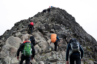 A group climbs up a steep, rocky mountain face. Two climbers await them at the peak on a cold, gray day.