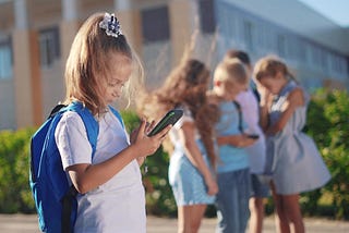 A young girl reads her cellphone alone as a group of children crowd around another cellphone behind her.