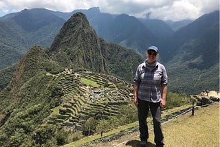 Nicole standing with blue cap, gray shirt, black pants. Background with green mountains and Machu Picchu.