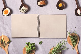 A blank recipe book open, surrounded by wooden spoons holding cheeses, eggs, herbs, garlic, and other ingredients.