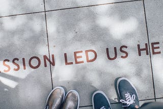 Looking down at two shoes on sun dappled concrete with the words “Passion led us here”.