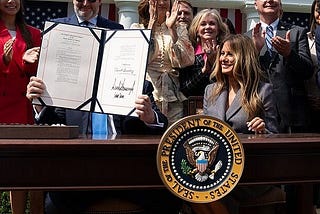 A picture showing President Donald Trump and First Lady Melania Trump participating in a bill signing ceremony for the Take it Down Act, Monday, May 19, 2025. Donald Trump is holding the bill showing their signautres while Melania is stting next to him, smiling.