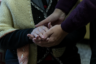 A pair of hands gently holds an elderly person’s hands wrapped in a cozy, knitted cardigan.