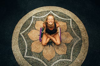 Yoga & Eastern Influence on Holistic Healthcare, photo of a young women, very flexible, sitting in a lotus position, on a lotus mandala designed on a floor.