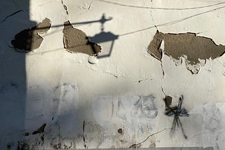 A photo of a cement wall, white paint peeling, graffiti barely covered with another coat of mismatched white. Morning sun shines on the wall, creating the shadow of a street lamp in the upper left corner.