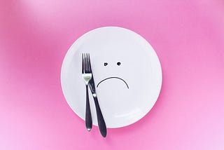 A white plate with a sad face and a fork and a knife over a pink background