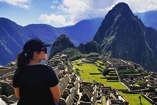 A woman wearing a black outfit and cap sits on a ledge overlooking the ancient Incan city of Machu Picchu, surrounded by towering green mountains under a partly cloudy sky.
