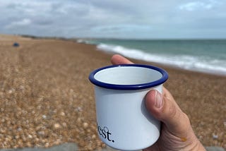 A hand holding a white cup with a blue rim on a pebble beach beside a calm sea.