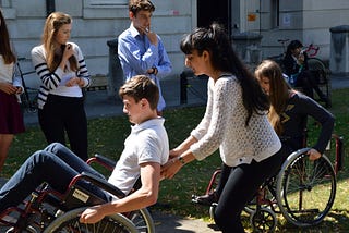 Young man in the wheelchair is looking a little scared as he tilts back in the wheelchair. His team mate is behind him to help 'spot' him. She looks equally anxious.