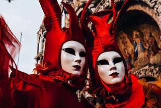 A pair of figures at a carnival, wearing elaborate red costumes and unsettling, almost featureless white masks