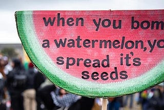 Photograph of a group of people at a Palestine protest with large sign in the shape of a slice of watermelon that reads “when you bomb a watermelon, you spread its seeds.”