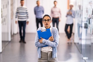 Woman standing in a hall, clutching a clipboard and looking worried.