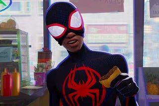 Spider-man Miles Morales stands in a black spider suit with a red spider logo on the front with the mouth part of his mask pulled up holding what appears to be a burrito maybe? Background is a bodega, which he is inside.