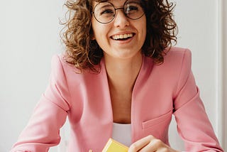 Woman in an office wearing glasses and a pink blazer holding sticky notes while looking up with a big smile on her face