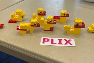 A flock of LEGO ducks sit on a table with a sign that says “PLIX”