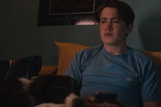 A scene of the character Nick Nelson in Netflix show Heartstopper. He is a white teenage boy with reddish hair wearing a blue shirt. He’s sitting on his bed, holding his phone and looking at nowhere, and his face expression is kinda thoughtful and worried. His dog Nellie, that has brown and white fur, is resting her head in his legs.