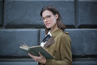 Serious adult woman wearing glasses in a smart shirt and necktie in a formal coat with open book on street against facade of old building in university. She’s looking inquisitively at the camera