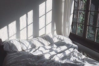 Sunlight streaming in the window, casting shadows on an unmade bed with white sheets.