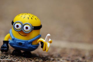 Close-up of Stewart the Minion figurine, a little smiling, bespectacled, yellow balding fellow with blue overalls, holding a banana against an outdoor dirt background.
