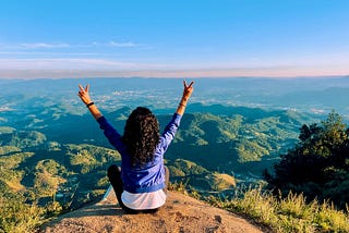 A woman sitting on a high hill with arms outstretched.