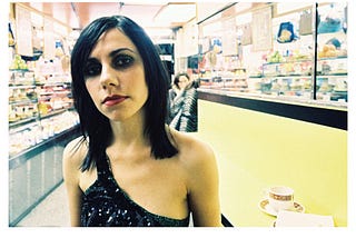 PJ Harvey “This Mess We’re In — Demo” single cover art; photo of Harvey in a black one-strap-sleeved black sequined top in a convenience store, white border with artist name on top and “STORIES FROM THE CITY, STORIES FROM THE SEA — DEMOS” text at bottom