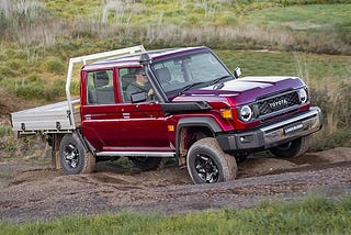 Metallic red 2024 Toyota Landcruiser VDJL79 series with black wheel crossing a rutted track. The wheels are articulated at opposing angles to show how capable the truck is off road. Behind the track is a grassy field.