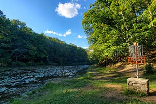 A disc golf basket sits near the edge of a lily pad-covered pond, surrounded by lush green trees under a bright blue sky with scattered clouds. The basket is mounted on a stone platform, marking hole 15 on a scenic wooded course.