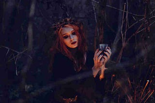 A pale red headed woman dressed in black holds a cup of a mysterious liquid in the middle of the forest