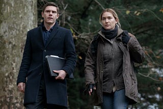 Two characters dressed for cold weather walk towards the camera in the woods. From left to right they are Colin Zabel played by Evan Peters and Mare Sheehan played by Kate Winslet.