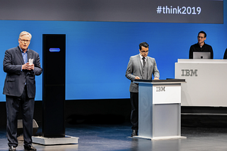 One person standing apart from others speaking, a tall black monolithic computer stand, another person behind a podium, and a group of people further back behind a low panel wall with hastag-think-2019 projected on a screen in the background