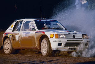 El Peugeot 205 Turbo 16 de rally avanza a toda velocidad sobre un terreno húmedo y fangoso, levantando una gran nube de agua y barro. La carrocería luce los logotipos de patrocinadores como Shell, Michelin y Lombard. Al volante, el legendario piloto finlandés Ari Vatanen, acompañado de su copiloto, maniobra con precisión en plena competición del Grupo B del Campeonato Mundial de Rally, demostrando su habilidad en condiciones extremas.