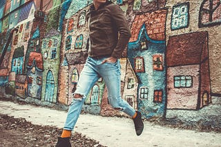 A stylish man in a casual outfit against a vibrant, colorful mural depicting whimsical houses. He seems to be walking on the air.