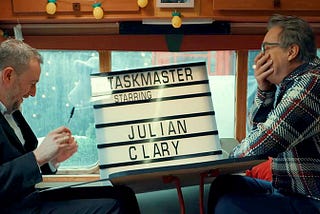 Julian Clary with his hand covering his mouth as he laughs, the table in the caravan destroyed