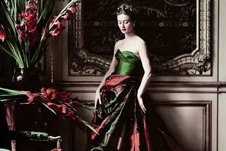 A model wearing a plaited up-do and a discreet wreath of red berries and green leaves poses in a green tafetta strapless bodice and a large skirt of dark green and berry red swagged at the waist. Gladioli in a vase match the berry red of the dress skirt and spill out above a green velvet seated chair. The model poses against a light brown panelled background with a dark chinosserie painting behind her. It’s all very country house ball.