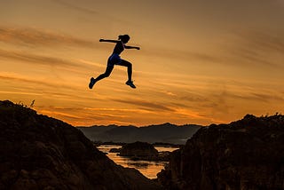 Young Woman In Sport Apparel Jumping Over A Cliff With Sunset In The Background.