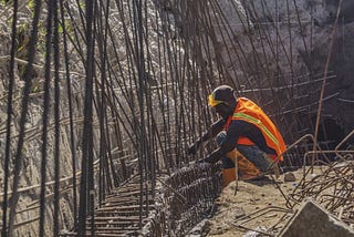Man wearing hard hat and orange safety vest is tying iron construction rods together.