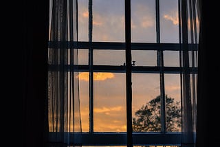 A view of the sky at dusk, with the silhouette of the window frame and sheer curtains in front.