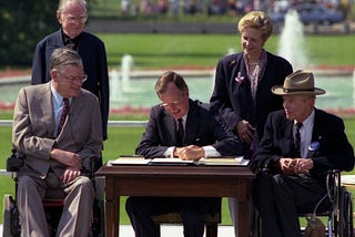 5 people in the image, Bush sitting in the middle and signing the ADA, 2 sitting on a wheelchair, and 2 standing at the back.