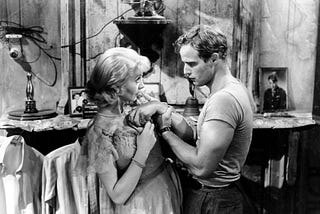 A black and white still of Stanley angrily grabbing Blanche’s arm, confronting her about what happened to Belle Reve while she is putting on perfume.