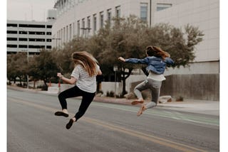 Two young women seen from behind, jumping and clicking their heels on a city street.