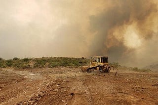 A bulldozer on a stretch of brown dirt, in front of a sky blotted out by smoke clouds.